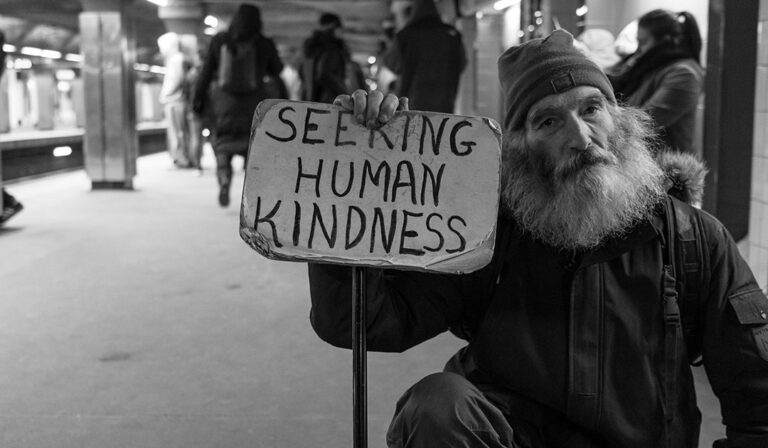 A man sitting on a sidewalk with a long shaggy beard holding a sign that says "seeking human kindness"