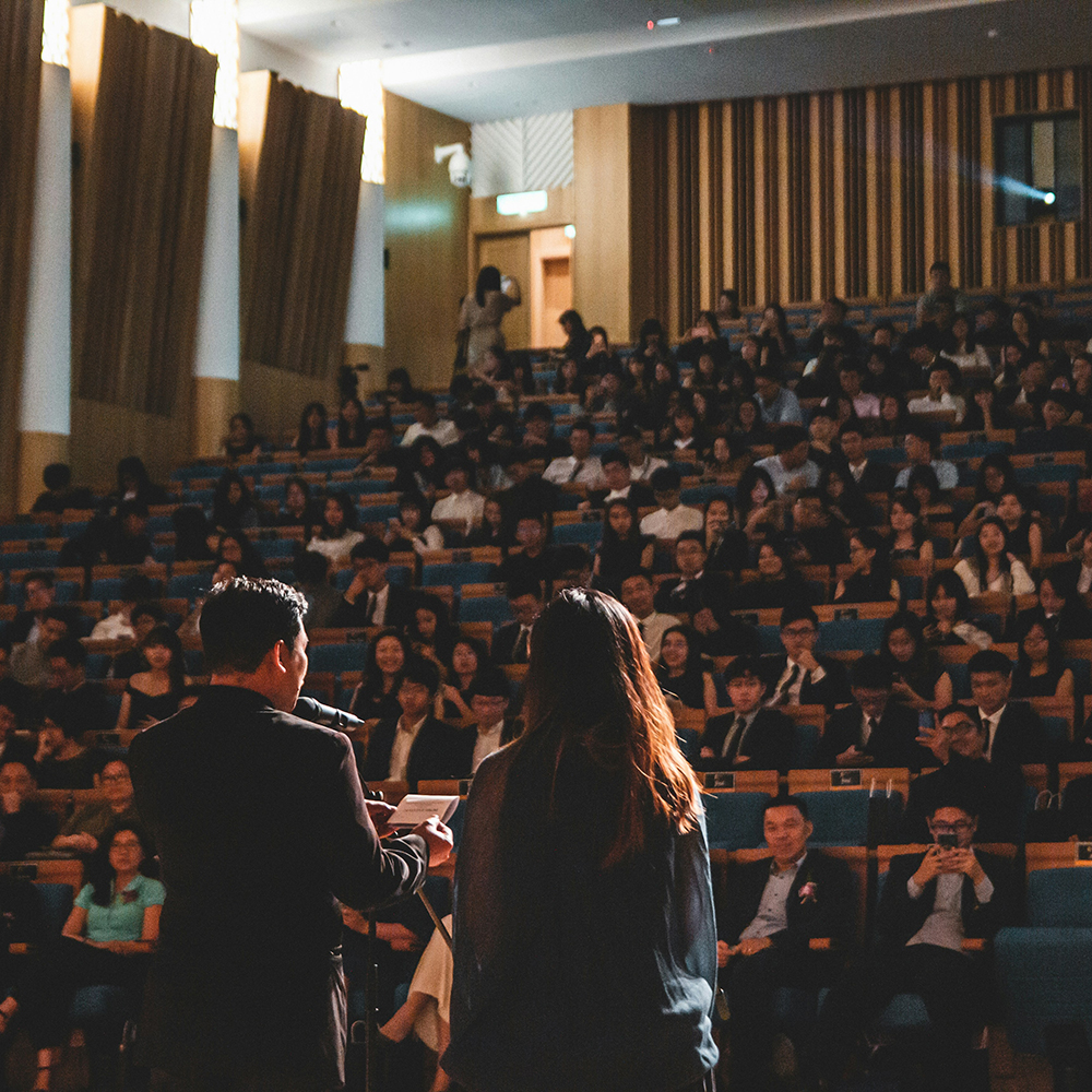 two people at a podium, from behind, speaking to an auditorium full of people